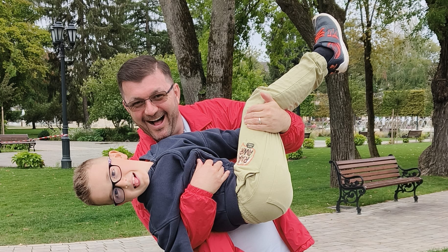 Ești mai sănătos decât tatăl tău? Tată în geacă roșie ridicându-și copilul într-un parc; zâmbet, energie și apropiere.